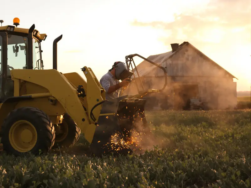 Welder repairing a loader bucket on a yellow tractor in a field at sunset, old barn behind