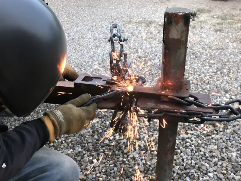 Close-up of a welder in a helmet and gloves welding a rusted trailer tongue and hitch on a gravel driveway, sparks flying